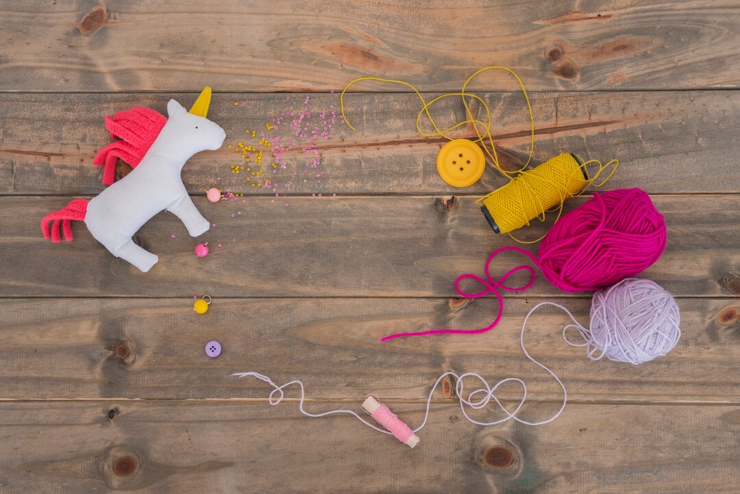 rag-unicorn-horse-with-yarn-pink-purple-spool-with-thread-button-wooden-desk_23-2148137400